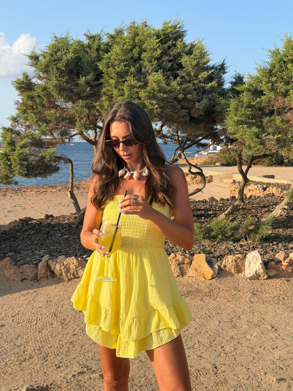 A woman wearing a bright yellow ruffled mini dress and sunglasses stands outdoors on a sandy beach-like area with trees, rocks, and blue water in the background, holding a drink.