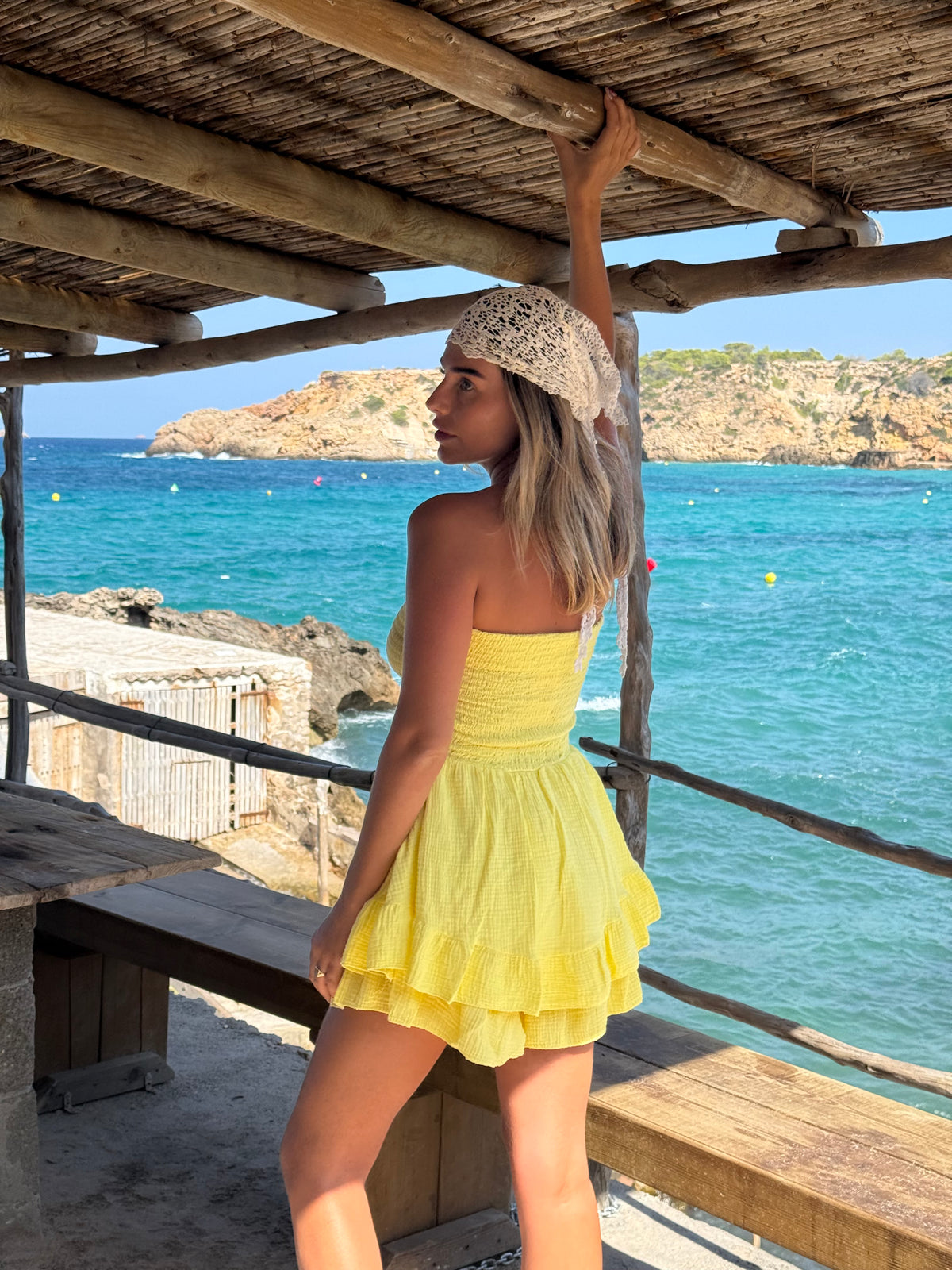 A woman wearing a bright yellow strapless ruffled mini dress and a white lace headscarf stands on a wooden beachside platform with a turquoise ocean and rocky coastline in the background.